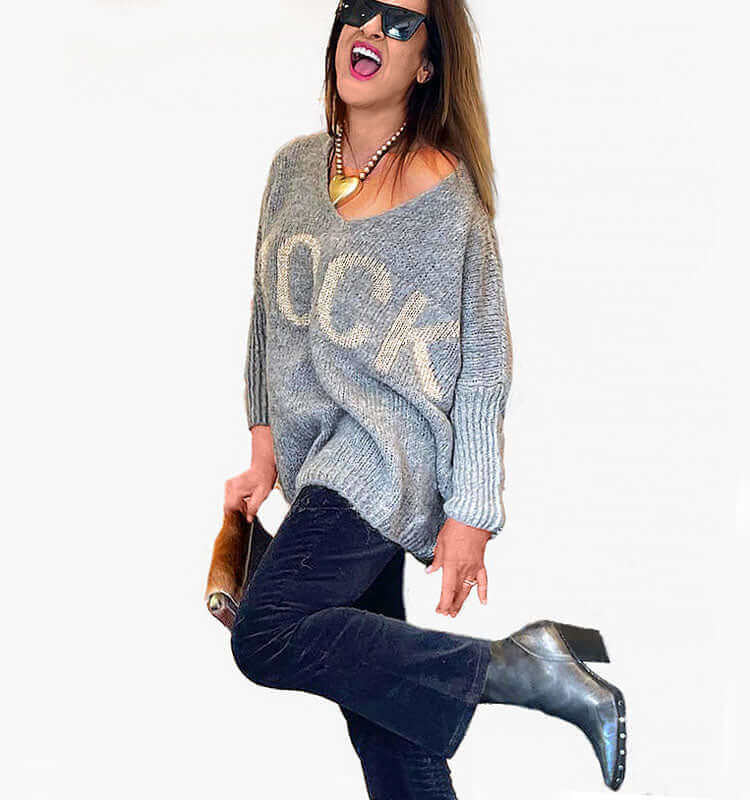 Woman in stylish oversized sweater and black jeans, posing playfully with sunglasses and ankle boots.