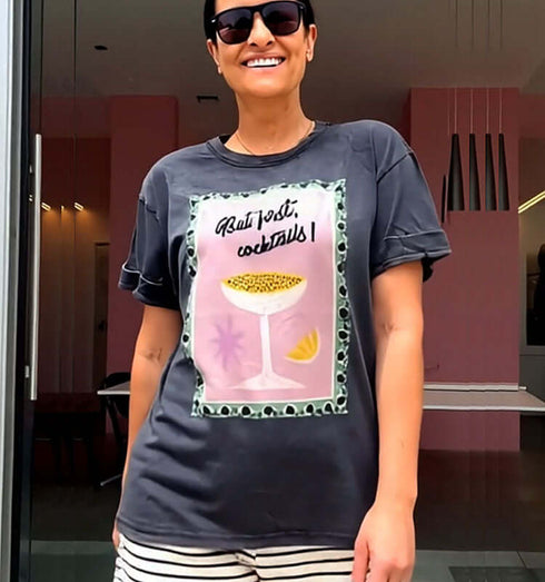 אישה עם טי-שירט אפורה וכחולה עם הדפס כוס קוקטייל, משקפי שמש ושוליים מפוספסים.