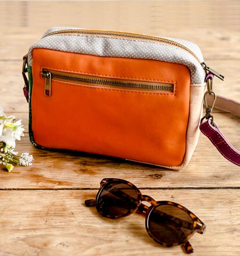 תיק צד מעור נפה בגווני תכלת, כתום עם רצועת כתף סגולה, ליד אובייקט שחור של משקפי שמש.