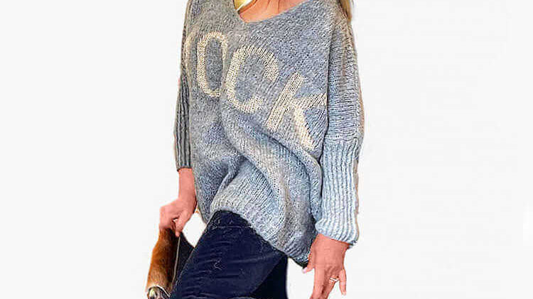 Woman in stylish oversized sweater and black jeans, posing playfully with sunglasses and ankle boots.