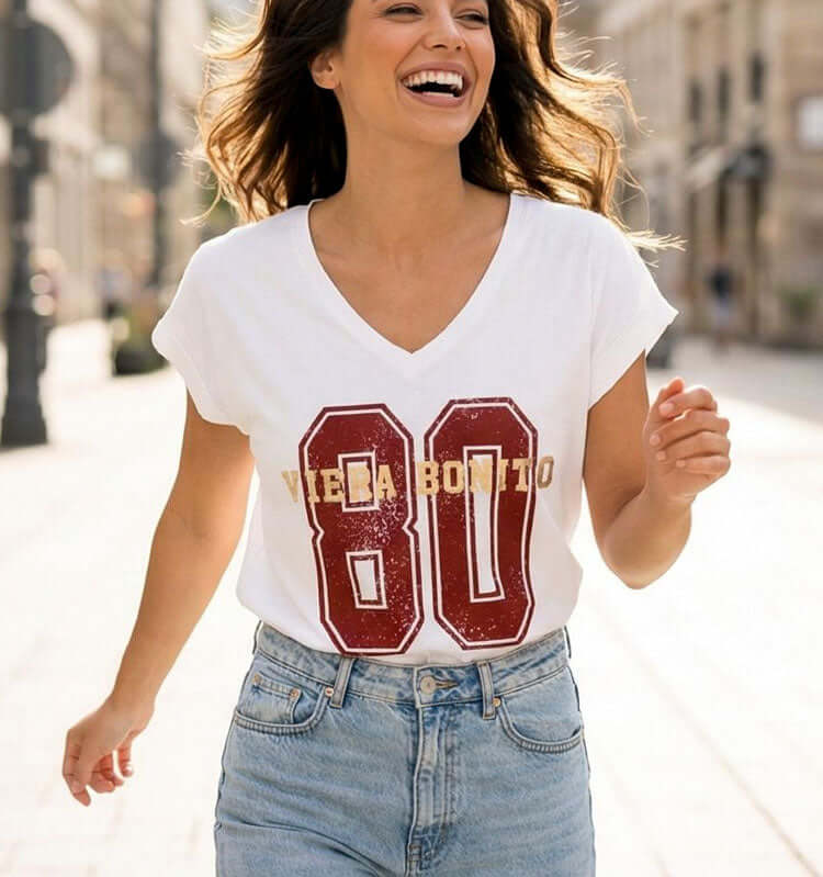 טישרט כותנה עם צווארון וי רחב בגוון לבן עם הדפ 80 בגוון אדום, לבושה על אישה מחייכת ברחוב.